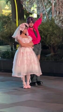 Two people stand on a paved surface outdoors in front of a building with greenery nearby. The first person is a young person dressed in a pink and white lolita-style dress with a matching bow and veil headpiece, white patterned tights, and pink shoes. They stand with their arms crossed and appear to be looking off to the side. The second person, with short, dyed purple hair, wears a bright pink button-down shirt, a black tie, dark jeans, and black shoes. This person moves in a manner that could be interpreted as dancing, first raising an arm, then stepping around the first person. The background features architectural elements, including an illuminated sign and a metallic-looking sculpture or decoration. The overall lighting suggests an outdoor setting during daytime or early evening.