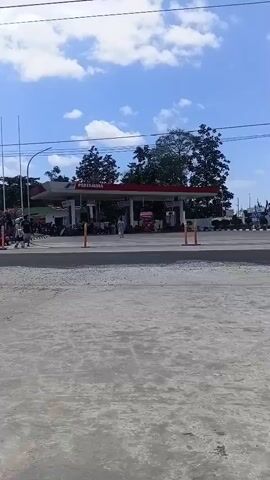 The video shows a Pertamina gas station under a partly cloudy sky. There are several motorcycles parked and in motion, with some people standing around the gas station. A white car is visible on the left side of the screen. The road in front of the gas station appears to be unpaved. There are trees and power lines in the background.