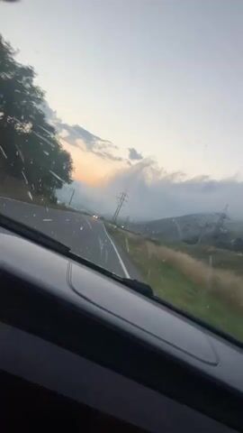 The video shows a view from inside a car as it drives down a road. The sky is overcast with clouds and a hint of sunset. There are trees on the left side of the road and rolling hills with power lines on the right. The road ahead appears to be empty except for a few distant car headlights. The car's dashboard is visible in the foreground. The windshield has water droplets on it, suggesting it might be raining or has recently rained.