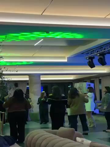 The video shows a group of women dancing in a brightly lit room with a modern ceiling. The ceiling features recessed lighting and stage lights that alternate between green and purple. There is stage equipment, like speakers, visible near the ceiling. The women are dressed in casual clothing and appear to be enjoying themselves. The room has a neutral color palette with beige furniture visible in the foreground and large windows in the background. The camera pans around the room.