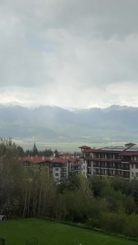 The video shows a view of a mountainous landscape on an overcast day. In the foreground, there are several multi-story buildings with red-tiled roofs surrounded by trees and green grass. Beyond the buildings, a valley stretches out towards a range of snow-capped mountains under a cloudy sky. The camera zooms in slightly on the mountain range and then focuses on a dirty window, before refocusing on the mountainous landscape.