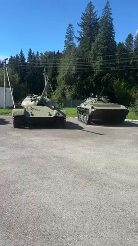 The video shows an outdoor scene with three military vehicles parked on an asphalt surface. In the foreground, a green tank is prominently displayed, facing forward. To its right, a camouflaged armored personnel carrier is also visible. In the background, a line of trees forms a natural backdrop, with a clear blue sky overhead. The overall impression is that of a military display or museum exhibit.