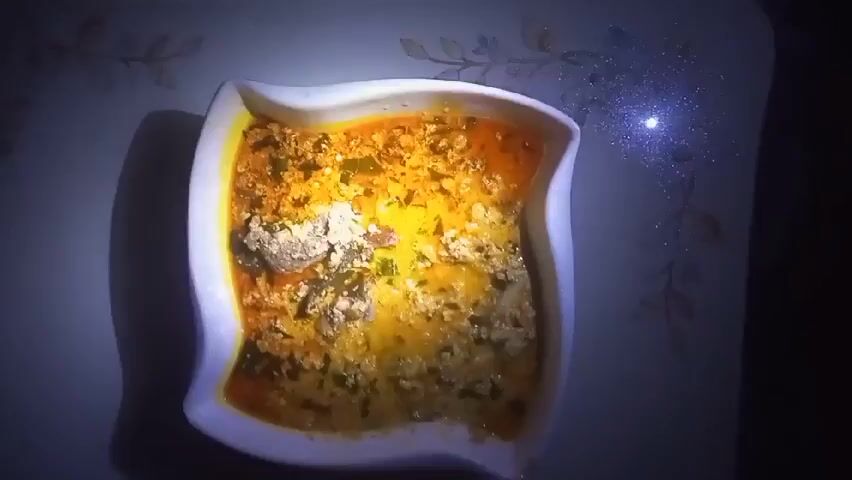 A still overhead shot shows a square, white bowl with a wavy rim filled with soup. The soup is a mix of orange, yellow, and green, and appears to contain chunks of meat or vegetables. The bowl sits on a table with a patterned surface. The lighting is somewhat dim, with a spot of light reflecting off the table near the top right corner.