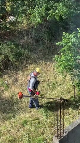 The video shows a man wearing a yellow cap, safety vest, and red gloves using a gas-powered string trimmer to cut grass and weeds on a small grassy incline. The man works methodically along the ground near a short concrete wall with exposed rebar. The surrounding environment includes trees, dense vegetation, and patches of dry grass.