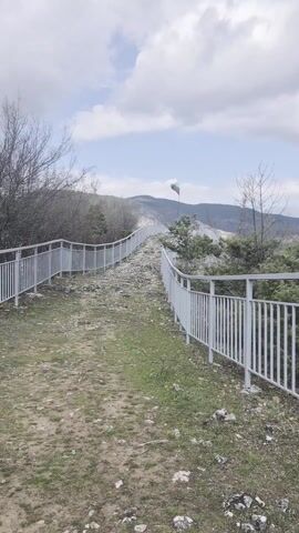 The video shows a path leading up a rocky hill, flanked by a white metal fence on either side. The path is dirt and grass-covered. At the top of the hill, a Bulgarian flag is waving. In the background, there are mountains under a sky with scattered clouds.  There are some trees and bare branches along the path. The video is taken from a static position looking upward along the path.