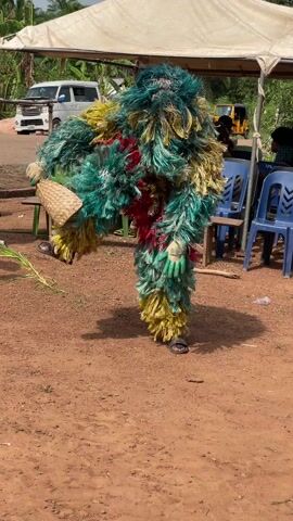 A brightly colored masquerade dancer in a fuzzy green, yellow, and red costume is performing outdoors on reddish-brown dirt. The dancer wears sandals, green gloves, and a woven conical hat in one hand, and takes exaggerated steps and arm movements to the accompaniment of drum music. In the background, other masked dancers, people seated under a tent, parked cars, and foliage are visible.