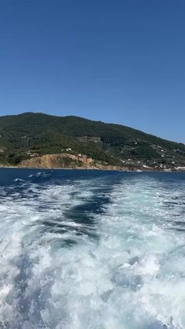 This is a first-person perspective video taken from the back of a boat moving through the sea. The focus is on the churning white wake left by the boat, which creates dynamic patterns in the deep blue water. In the distance, a lush green, mountainous island rises from the sea. White buildings are visible scattered across the slopes of the island, suggesting a coastal village or town. The sky above is a clear, vibrant blue, indicating a sunny and pleasant day. The scene is set in Skopelos, Greece, a location known for its beautiful natural landscapes and picturesque villages. The video captures a sense of travel and exploration, with the viewer experiencing the journey as the boat moves away from the island. The overall mood is serene and idyllic, highlighting the beauty of the Mediterranean coastline. There is no visible text in the video.