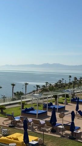 The video shows a scenic view of a resort with a swimming pool, lounge chairs, and cabanas. The resort is situated by the sea, with palm trees lining the coast. In the background, mountains can be seen across the water. The sky is clear and blue. The resort building is white and modern in design. The lounge chairs are blue, and the cabanas have blue cushions. The overall atmosphere is peaceful and serene.