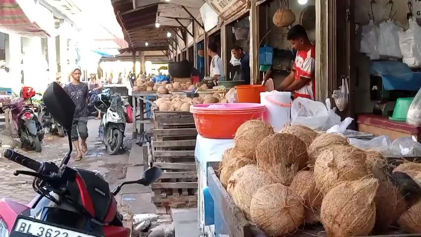 This is a horizontal, eye-level shot of a bustling market scene, likely in Southeast Asia. The foreground is dominated by a display of coconuts, piled high in wooden crates. To the left, a red motorcycle is parked, with its front wheel and handlebars clearly visible. A person walks from left to right in the mid-ground, carrying a large black object over their shoulder. Further back, several more motorcycles are parked. On the right side of the frame, a market stall is set up, with several people working. One man in a red and white striped shirt is actively preparing coconuts, while others appear to be tending to their stalls. The stalls are covered by awnings, and the overall environment suggests a warm, sunny day. The aisle between the stalls and the parked motorcycles is paved, with some debris on the ground. The focus is on the marketplace activity, highlighting the abundance of coconuts and the daily commerce taking place.