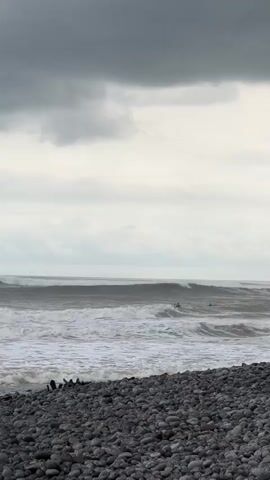 A vertical video shows a rocky beach in the foreground and the ocean in the background. Several waves are breaking in the ocean, and a few surfers are visible in the water. The sky is overcast with gray clouds.
