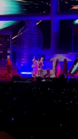 The video shows a live performance on a stage with a large screen in the background displaying colorful abstract graphics. There is a prominent graphic of a clear plastic bottle on the right side of the screen. On the stage, there is a female singer in the center, wearing a light-colored outfit, holding a microphone and singing. Around her are several dancers in matching outfits, some in orange and some in white, performing synchronized choreography. They are arranged in a semi-circle around the singer. The stage is lit with bright blue and red lights, creating a vibrant atmosphere. In the foreground, a dark area with a crowd of people with their phones raised can be seen, indicating an audience.
