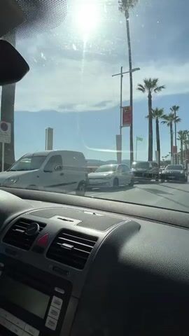 The video shows a view from inside a car driving along a road lined with palm trees. The sun is bright, suggesting it is daytime, and the sky is blue with some scattered clouds, indicating pleasant weather. The scene appears to be a coastal town, possibly a promenade, with buildings and more palm trees visible on the right.

Several cars are on the road, and some are parked along the side. A white car is visible on the left side of the road, and a darker van is to its right. Further down the road, there are more cars and people walking along the sidewalk. A green flag is flying from a pole. A red and white sign is attached to a lamppost with some text on it. A red car is also visible, as is a person riding a bicycle. The sea is visible in the background on the left, with a few people near the water. The overall atmosphere is relaxed and suggests a vacation or a leisurely drive. The interior of the car is visible in the foreground, with the dashboard, air vents, and radio display. There are no discernible emotions conveyed, and the focus is on the external scenery and the movement of the car.
