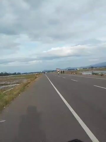 A motorcycle is traveling on a paved road with a white line down the center. To the left of the road, there are fields that appear to be flooded, possibly rice paddies. To the right of the road, there are more flooded fields and some trees in the distance. The sky is cloudy. Several people are visible on the right side of the road, some standing and some on motorcycles. One person is standing on the side of the road and appears to be looking at the motorcycle that is filming the video. There are also a few vehicles in the distance on the road.