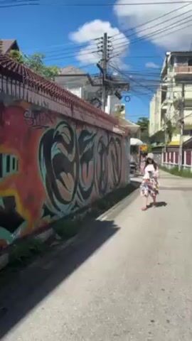 A young woman walks on the side of a street with a colorful graffiti wall on her left. The wall is large and features abstract, organic shapes and lettering. The woman carries a drink in her left hand and a phone in her right. She is wearing a white top, a light pink sweater, a floral skirt, and a light pink bucket hat. The street is narrow, lined with buildings on both sides, with tangled wires overhead. The sky is blue with a few clouds.