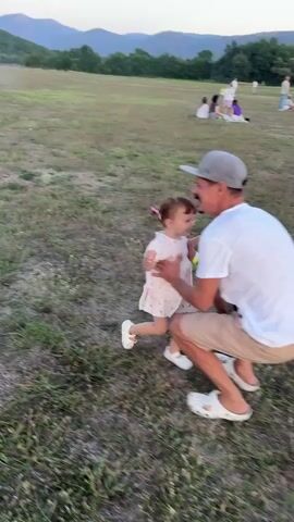 The video shows a father interacting with his young daughter in an outdoor grassy field. In the first frame, the father, wearing a white t-shirt, beige shorts, a baseball cap, and white Crocs, is crouching and holding the girl, who is wearing a light-colored dress. The girl has her arms around the father's neck. In the second frame, the father is standing and lifting the girl into the air, both smiling. In the third and fourth frames, the father is holding the girl on his shoulders, facing away from the camera, with the girl looking over his shoulder. In the background, there are hills and trees, a clear sky, and other people gathered in the field, some sitting and some standing, suggesting an outdoor event. The ground is covered with dry grass and some scattered purple flowers.
