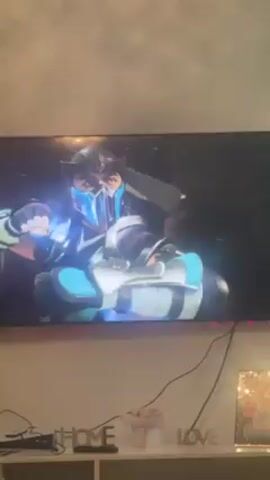 A video shows someone playing Mortal Kombat on a television. The game shows two characters fighting in a purple-lit arena. The words “ROUND 1”, “FIGHT”, and the character's health bars are visible on the screen.  Below the television is a shelf with decorative objects, including a sign that reads “HOME LOVE”. The television is surrounded by colorful LED strip lights.