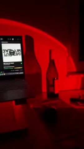The video shows a desk bathed in red light. There is a laptop on the left side of the desk, with a music streaming service open, displaying the title "GET OUT MY HEAD." Next to the laptop is a wine bottle and a large glass bottle, presumably empty. A mouse and a mousepad are visible in the foreground. The red light casts strong shadows of the bottles onto the wall behind the desk. The overall aesthetic is moody and dramatic due to the intense red lighting.