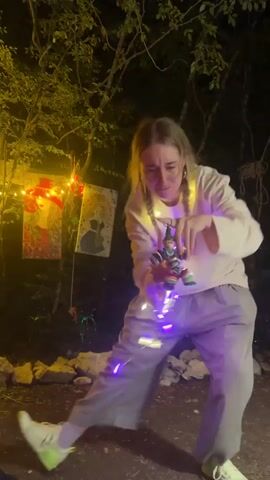 A young woman with blonde braided hair, wearing a white sweatshirt and grey pants, stands in a dimly lit outdoor area decorated with string lights. She is holding a small, colorful, cylindrical object with some green elements, possibly a toy or a craft item. She appears to be examining it closely and speaking. Two framed pictures hang on a dark background behind her, illuminated by the lights. The ground is rocky and uneven. The overall atmosphere suggests a nighttime gathering or event.