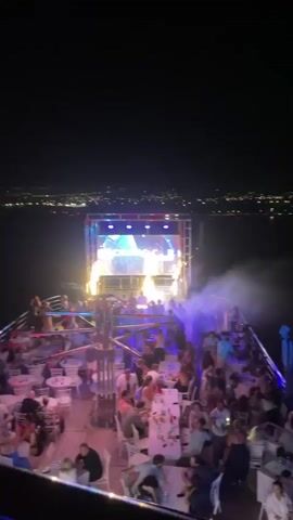 This media file depicts a vibrant night scene at an outdoor event, likely a party or club night, taking place in Yeroskipou, Cyprus. The primary subject is a large crowd of people enjoying themselves in a lively atmosphere.

The scene is set on a multi-level platform, possibly a boat or a rooftop venue, judging by the railing and elevated perspective. In the background, the distant lights of a city or town are visible against a dark night sky, suggesting the event is overlooking a populated area.

A prominent feature of the scene is a large LED screen mounted on a metallic truss structure. This screen displays dynamic visuals, cycling through different graphics and colors. At various points, the screen clearly shows the text "OCEAN Vision" in a stylish font. The visuals are accompanied by bright, colorful lighting that bathes the venue in shades of blue, green, and pink, creating a festive and energetic ambiance.

The activity taking place is clearly a celebration or entertainment event. People are gathered, with many standing and dancing, while others are seated at tables, presumably enjoying drinks and conversation. There is a notable presence of fog or smoke machines, which add to the atmospheric lighting and visual effects. Sparks are also seen erupting from the stage area at one point, indicating a performance or special effect.

The overall mood conveyed is one of excitement, fun, and revelry. The time of day is night, and the weather appears clear, allowing for the distant city lights to be visible. The location cues point to Yeroskipou, Cyprus, suggesting a coastal or Mediterranean setting. The people are dressed casually, fitting for a night of dancing and socializing.