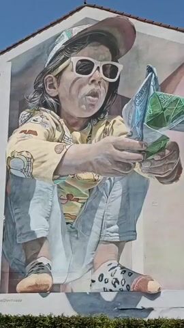 The video pans across a large mural painted on the side of a building. The mural depicts a young child wearing sunglasses, a cap, and a yellow shirt with a cartoon design. The child is crouched down and appears to be holding and manipulating a folded piece of paper or packaging, possibly a wrapper, with both hands. The child's mouth is open as if in mid-action, and there are remnants of something white around their mouth. The mural utilizes realistic rendering and shading to create a three-dimensional effect, with shadows cast on the wall below. The background of the mural shows architectural elements. The video then shifts to reveal the real-world environment surrounding the mural, showing adjacent buildings, windows with laundry hanging out to dry, and a clear blue sky. The bottom of the frame shows some green foliage. A logo and text are visible at the lower right of the mural.