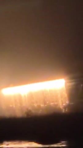 This is a short video clip of a bright, rectangular light source that appears to be in motion, possibly descending or moving horizontally. The light is intensely orange and yellow, with a glowing effect that causes it to bleed into the surrounding environment. The background is hazy and dark, with some suggestions of structures or terrain below the light. The camera appears to be moving as well, possibly in a vehicle, as indicated by slight motion blur and a horizontal perspective. The overall impression is of a distant, luminous object observed in low-light conditions, possibly at night or during dusk/dawn.