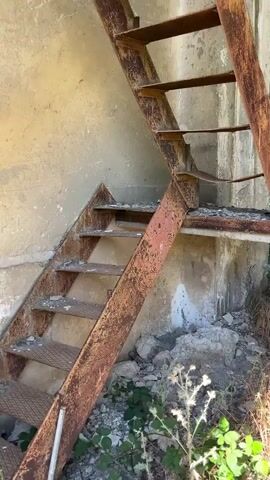 The video pans through a dilapidated concrete structure. The first shot shows a rusty metal staircase against a concrete wall, with debris and vegetation at its base. The subsequent shots reveal the interior of the building, with open doorways, exposed brickwork, and overgrown plants. The camera then looks upwards, showing the decaying structure with open ceilings and exposed beams, with sunlight filtering through. The final shots continue to show the upward view of the damaged building against a clear blue sky, with greenery visible through the openings.