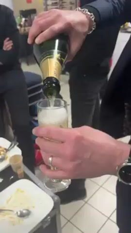 A person is pouring champagne into a champagne glass. The individual wears a dark blue blazer, a silver ring, and a silver watch. In the background, two people stand, and one sits on a beige couch. The scene takes place in a backstage or dressing room area with finished plates of food and glasses on a table. The floor is tiled. There is a brand of champagne, "Hoguet", visible on the bottle.