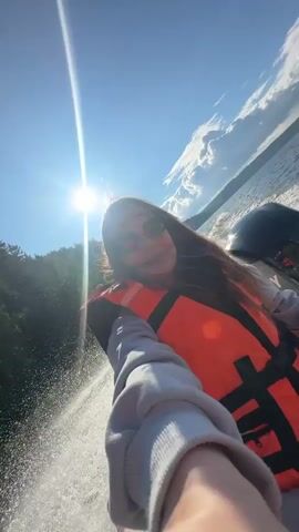 A young woman with long brown hair and sunglasses is riding on a boat. She is wearing an orange life jacket over a grey hoodie and is taking a selfie. The boat is moving fast, creating a spray of water behind it. The sun is shining brightly in a blue sky with some clouds. In the background, there is a body of water and a tree-covered hillside. The woman smiles and makes a peace sign with her fingers.