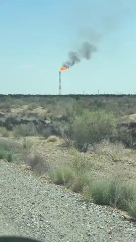 The video shows a tall flare stack with a large flame and dark smoke emanating from the top. The flare stack is situated in a flat, arid landscape with sparse vegetation, including bushes and dry grasses. In the background, more industrial structures and power lines are visible. The camera appears to be in motion, possibly from a vehicle, as indicated by the gravel road in the foreground and the slightly blurred vegetation. The sky is clear and blue. The overall scene suggests an industrial site, likely an oil or gas processing facility, in a remote or undeveloped area.