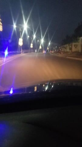 The camera is stationary inside a vehicle, looking out the side window onto a city street at night. There are a few cars with headlights on the street. Streetlights are visible with the light refracting.