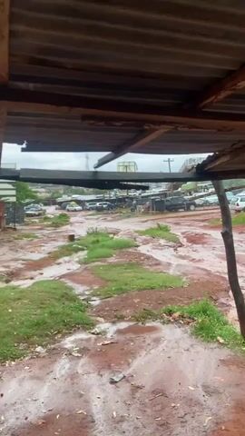 The video captures a static wide shot of a muddy, unpaved outdoor area in Auchi, Nigeria, likely after a recent rainfall. The foreground is dominated by red-brown earth, heavily saturated with water, forming numerous puddles and small channels where water has flowed. Patches of green grass are interspersed throughout the muddy ground.

Overhead, a corrugated metal roof, supported by rough wooden beams, extends into the frame, providing shelter from which the scene is viewed. In the midground and background, various vehicles, including cars, trucks, and SUVs, are parked on the wet, unpaved surface. Some dark plastic containers, possibly water tanks, are visible near certain vehicles. The overall area appears to be an informal parking lot or a section of a market.

The sky in the distance is overcast, contributing to the muted lighting. No people are visible, and no specific activity is taking place. The scene conveys a sense of post-rain quietude in a somewhat rugged and undeveloped setting. No visible text is present in the video.