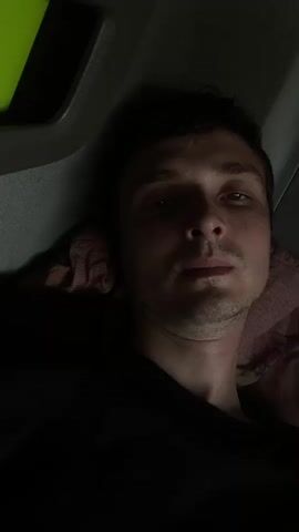 This media file is a close-up, selfie-style video of a young man lying down and looking directly at the camera. He has short, dark hair and appears to be in a dimly lit environment, possibly a vehicle or a confined space. His expression is neutral, with his eyes open and looking forward. The lighting casts shadows on his face, highlighting his features. He is wearing a black t-shirt. Behind him, a patterned fabric, likely a pillow or blanket, is visible. There are no visible signs of text in the media file. The video is taken indoors, suggesting an interior setting rather than an outdoor scene. The overall mood is subdued due to the low lighting.
