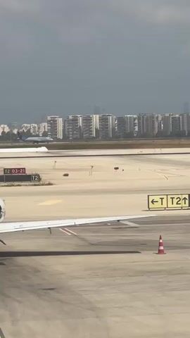 The video shows an airport tarmac. In the foreground, the wing of a parked airplane is visible on the right. Further back, a white airplane is seen landing on the runway, kicking up dust. Beyond the runway, there are several apartment buildings and cityscapes in the background. The sky is overcast.