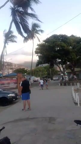 The video shows a street scene with palm trees swaying in the wind. In the foreground, a person is sitting on a motorcycle, their hand on the handlebar. To the left, a dark-colored car is parked. Further down the street, several people are walking and standing. A man in a black shirt and blue shorts is facing away from the camera. To his right, a group of people are standing and walking. A white motorcycle with a rider wearing a white shirt is moving away from the camera down the street. In the background, there are buildings on a hillside and more trees. The sky is overcast with some clouds.