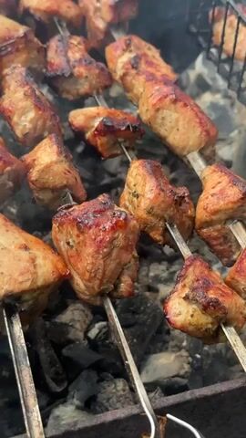 This is a close-up, vertical, eye-level shot of several skewers of meat being grilled over hot coals. The meat is cubed and appears to be pork, with a reddish-brown, slightly charred exterior and visible herbs and spices. The skewers are arranged on a metal grill rack. The background consists of glowing embers and ash, indicating an active barbecue. There is a slight haze of smoke rising from the coals. The lighting suggests it is daytime, likely outdoors, given the presence of a charcoal grill. The overall impression is one of a delicious and savory meal being prepared. There is no visible text in the media.