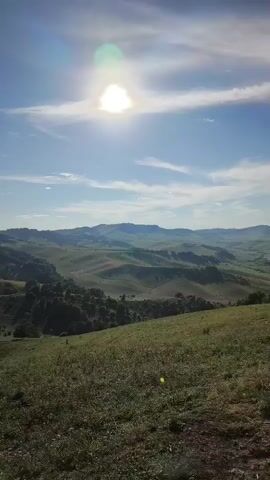 A video shows a panoramic view of a vast landscape. The sun shines brightly in the sky, casting a warm glow over rolling hills covered in lush green grass and scattered trees. In the distance, a wide river winds its way through a valley, flanked by dense forests and more rolling hills. The sky is a clear blue, with a few wispy clouds adding texture. The camera pans slowly across the scene, giving a sense of peace and tranquility. There are no people or man-made structures visible in the video.