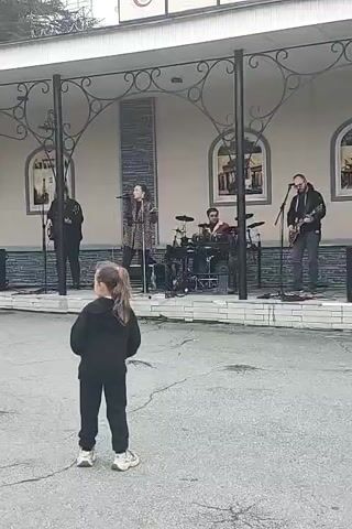 A young girl with her back to the camera, wearing a black hooded sweatshirt and pants, stands on asphalt. She is facing a band performing on a raised platform. The band consists of a female singer in the center, wearing a patterned jacket, and three male musicians: one playing a guitar to her left, another playing drums behind her, and a third playing a guitar to her right. The platform has a decorative black metal railing and a light-colored floor. The background features a building with arched windows containing images. The overall scene appears to be an outdoor event.