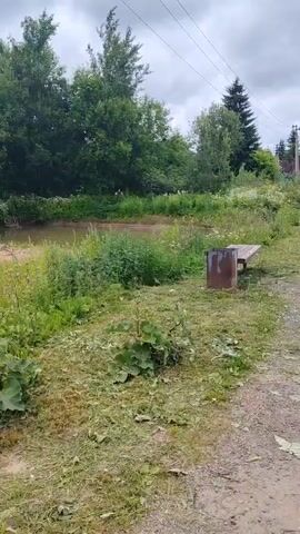 The video shows a view of a natural setting next to a body of water. In the foreground, there is freshly cut grass and some vegetation. To the right, there is a rustic wooden bench next to a rusty metal trash can. Behind the bench and trash can, tall green grass and weeds grow. In the middle ground, a calm body of water, possibly a river or pond, is visible. The water has a muddy or silty bottom, with some vegetation growing at its edge. The background consists of lush green trees and foliage, forming a dense treeline. The sky is overcast with clouds, suggesting a temperate or possibly humid day. There are no people or animals visible in the video. The scene appears to be an outdoor recreational area that has recently been maintained, with the cut grass indicating recent activity.