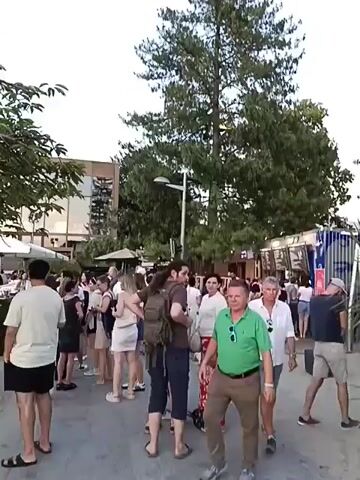 The video shows an outdoor gathering of people. In the foreground, a man in a green shirt walks towards the camera. Behind him, a crowd is milling around. There are food stands or kiosks set up, some with menus visible. A large tree dominates the background, and a modern building with large windows is also visible. People are dressed casually in warm-weather clothing. Some individuals are standing in lines, possibly waiting for food or drinks. The overall atmosphere appears to be relaxed and social, possibly at an outdoor market or event.