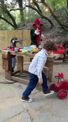 Outside on a stone patio in front of an artificial fairy house with a bright blue round door, a small boy wearing a light blue and white plaid shirt and dark blue jeans picks up a red and white striped candy cane and hands it to an elf dressed in green and red standing behind a wooden table covered with craft materials. A small girl with red hair and a red t-shirt stands beside the elf. A red toy car is on the stone patio next to the boy. Trees are in the background.