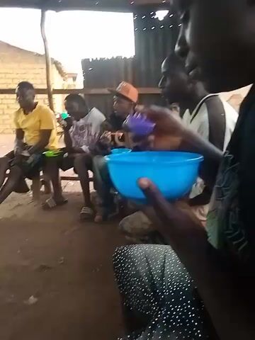 The video shows a group of people in what appears to be a small, outdoor eatery or gathering space. Several men are seated on benches or stools, eating from blue bowls, under a rudimentary shelter constructed with wooden posts and a corrugated metal roof.  A larger blue bowl sits in the foreground. Further inside the shelter, other people are gathered around a makeshift cooking area with an open fire. They appear to be preparing or serving food, with one person frying something in a pan over the fire. There are several containers nearby, including buckets and a red plastic bowl. In the background, outside the shelter, there are glimpses of buildings, trees, and possibly a vehicle, suggesting a village or small town setting. The ground appears to be dirt or sand.