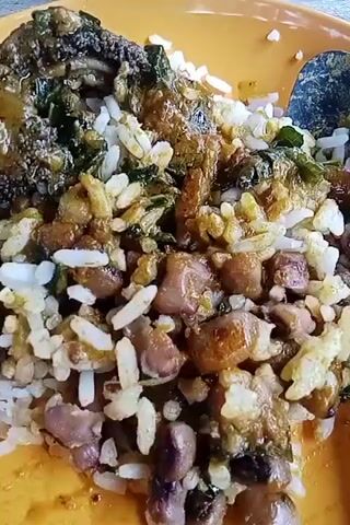 A plate of rice and beans topped with a green leafy vegetable-based dish. The rice is white. The beans are dark brown. The green leafy vegetable dish appears to have a sauce. The plate is yellow.  There is a bowl of what appears to be stew in the background. There is also a glass with dark liquid in it in the background. A dark grey object sits adjacent to the plate. A silver-colored eating utensil is next to the plate. A person says "local but it's sweet".