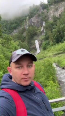 A man wearing a blue jacket, a red backpack, and a dark baseball cap is standing in front of a waterfall in a forest. He is looking at the camera with his mouth open in surprise. A stream is flowing next to him, and trees are growing on the hillside. The sky is overcast.