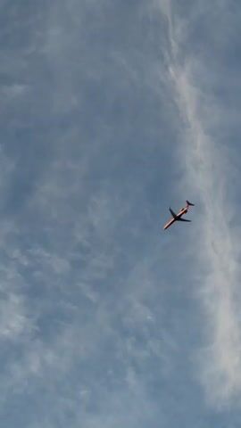 The video shows a small, orange airplane flying in a bright blue sky with scattered white clouds. The airplane is seen from below and appears to be a commercial jet with two engines under its wings. The camera is fixed on the airplane, and the view is slightly tilted. The airplane is flying from left to right across the frame and appears to be at a moderate altitude. The video is short, lasting only a few seconds, and there is no audio.