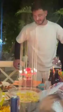 A person with glasses stands behind a table with several dishes and drinks on it. The table is outside and has a white, lattice-style trim. Behind the person, trees and string lights are visible. On the table, there's a cake with lighted candles. The person looks down at the cake, then to the side, smiles, and sticks out their tongue.