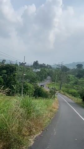 The video shows a car driving on a winding road. In the distance, there are several people standing near a motorcycle on the side of the road. A black van is parked nearby. The road is surrounded by green vegetation and trees. Power lines run along the left side of the road. The sky is partly cloudy.