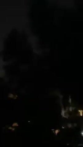 A dark, blurry video shows what appears to be trees and the faint outlines of buildings at night. A bright flash of light momentarily illuminates the scene, followed by a loud "meow" sound.