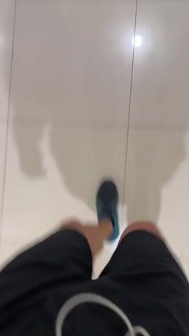 A first-person perspective video shows the legs and feet of an individual walking forward. The person is wearing black athletic shorts and dark running shoes with blue and green details. They are walking on a light-colored tiled floor, which has visible grout lines. Shadows, consistent with an overhead light source, are cast on the floor and move with the person's steps.