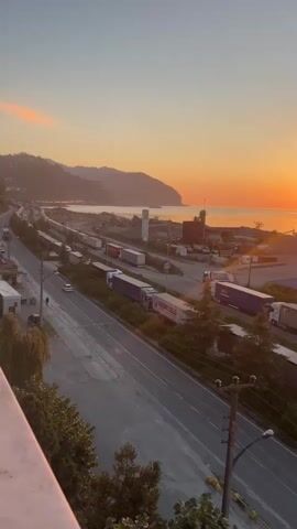 The video captures a wide shot of a coastal highway at sunset. The sun is setting over the ocean, casting an orange glow across the sky and reflecting on the water. A road runs parallel to the coast, with several lanes of traffic. A long line of parked semi-trucks is visible on the side of the road. In the background, there are hills, a port with industrial buildings, and a lighthouse. The overall scene depicts a busy transportation hub during a picturesque sunset.