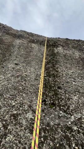 The video shows a person rappelling down a rock face using a yellow and red rope attached to a harness. The camera first focuses on the rope descending the gray rock face. Then, the perspective shifts to show the person's hands holding the rope and their climbing harness. The camera then pans out to reveal a view of the green valley below, with a dirt path winding through the grass. The final shot shows a wider view of the valley, with rock formations in the distance and mountains further beyond. The sky is overcast and gray.